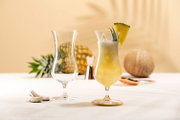 Vooraanzicht van twee elegante hurricane cocktailglazen, één leeg en één gevuld met een romige tropische ananas-kokosdrank, ijs en een schijfje ananas
