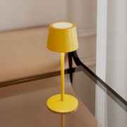 Gelbe, moderne Tischlampe mit konischem Schirm und rundem Fuß auf einem Glastisch neben einem beigen Sofa, aufgenommen aus leicht erhöhter schräger Perspektive