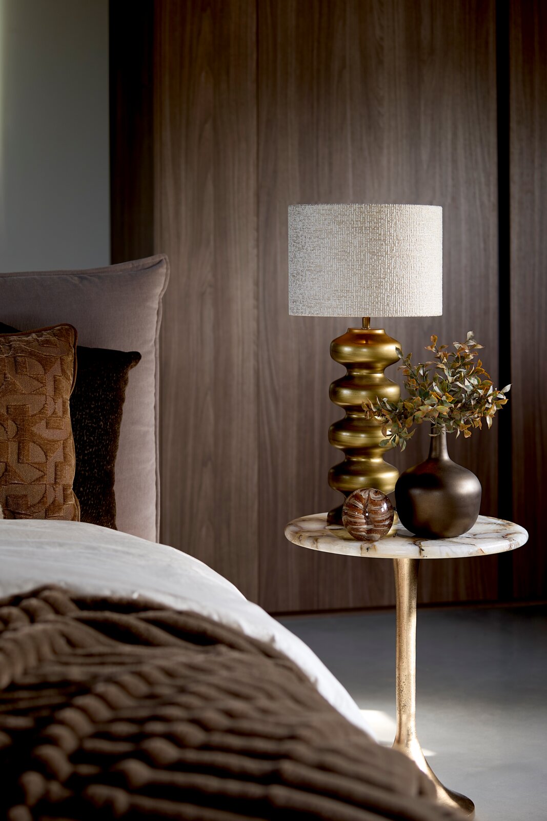Messingkleurige tafellamp met organisch gevormde voet en beige stoffen kap, van voren gefotografeerd op een marmeren bijzettafel naast een stijlvol opgemaakt bed.