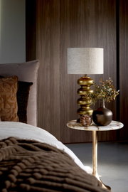 Messingkleurige tafellamp met organisch gevormde voet en beige stoffen kap, van voren gefotografeerd op een marmeren bijzettafel naast een stijlvol opgemaakt bed.