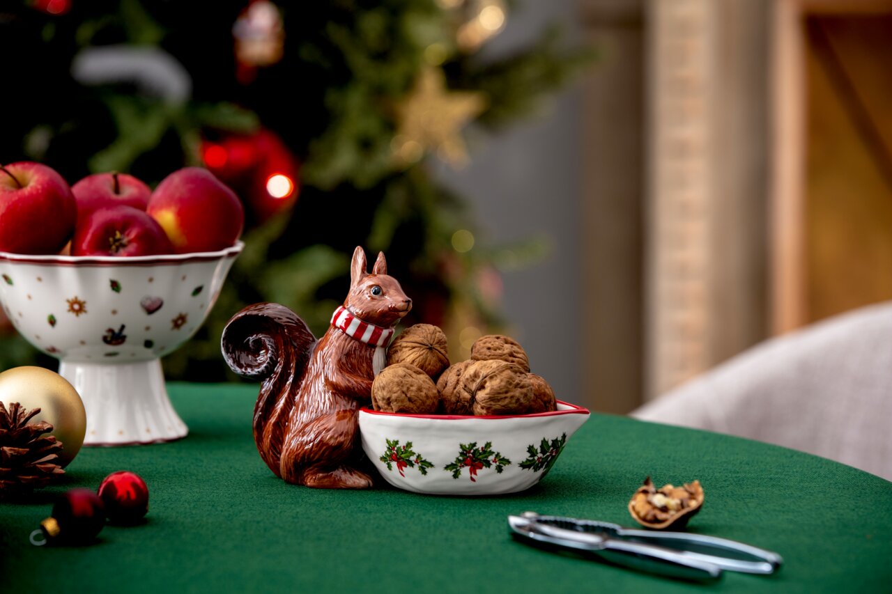 Keramik-Eichhörnchen mit Schal hält eine Schale mit Walnüssen, auf einem festlich gedeckten Tisch mit Weihnachtsdekoration, seitliche Perspektive.