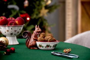 Keramik-Eichhörnchen mit Schal hält eine Schale mit Walnüssen, auf einem festlich gedeckten Tisch mit Weihnachtsdekoration, seitliche Perspektive.