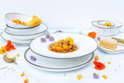 Weiß gesprenkeltes Porzellan-Geschirrset mit tiefem Pastateller, Schalen und Tellern mit braunem Rand, dekorativ angerichtet mit Speise und Blüten, fotografiert in leicht erhöhter Frontansicht.