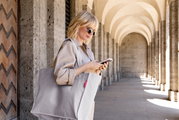 Frau mit Sonnenbrille trägt eine große hellgraue Stoff-Tote-Bag mit rotem Label über der Schulter und schaut auf ihr Smartphone, Seitenansicht in einer Arkadengang-Umgebung.
