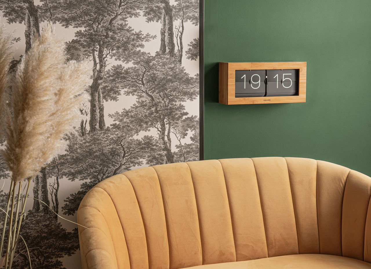 Wanduhr aus Bambus mit Klappzahlen, montiert an einer grünen Wand, im Hintergrund eine Tapete mit Baum-Muster und ein gelbes Sofa im Vordergrund, seitliche Perspektive.
