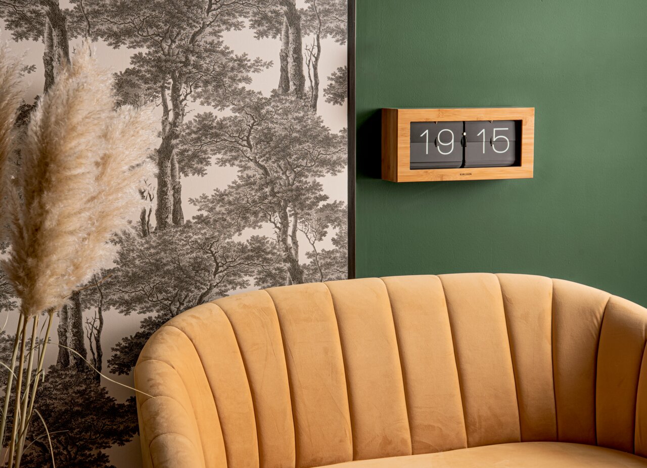 Bamboe wandklok met uitklapbare cijfers, bevestigd aan een groene muur, op de achtergrond behang met een boompatroon en op de voorgrond een gele bank, zijaanzicht.