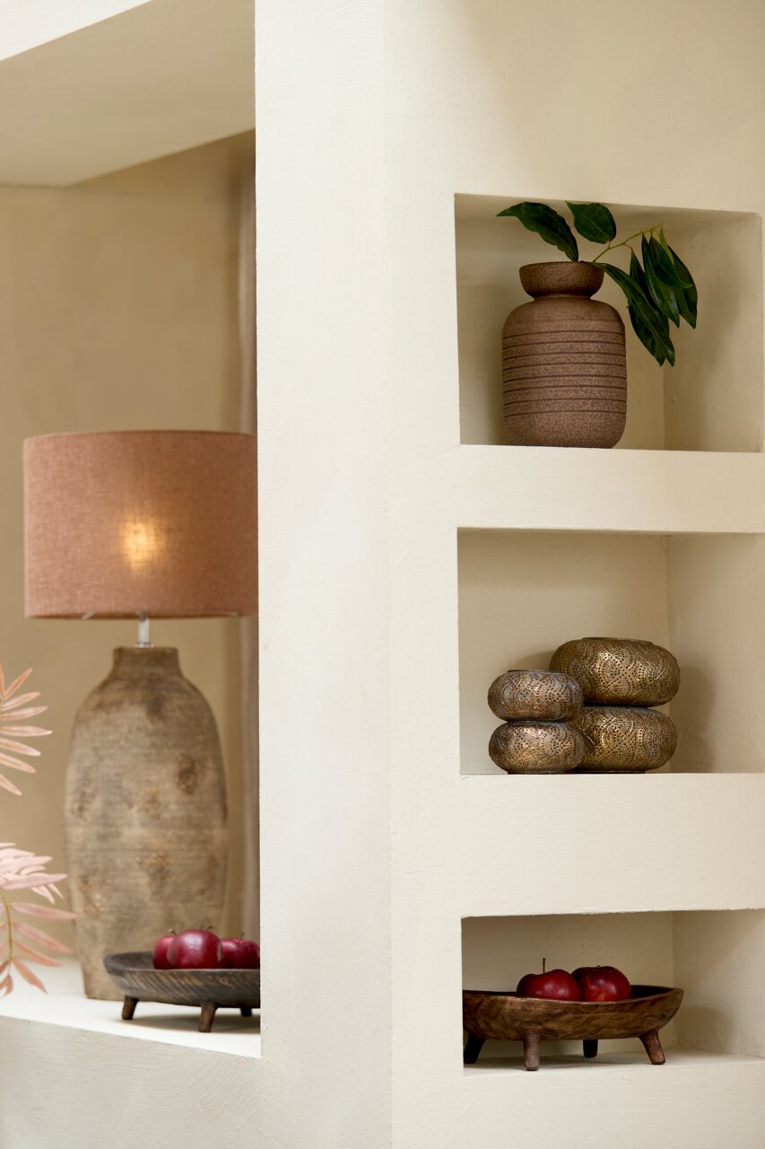 Ein stilvolles Windlicht in einem Regal, umgeben von dekorativen Elementen wie einer Vase mit Blättern und einer Tischlampe. Perspektive von vorne.