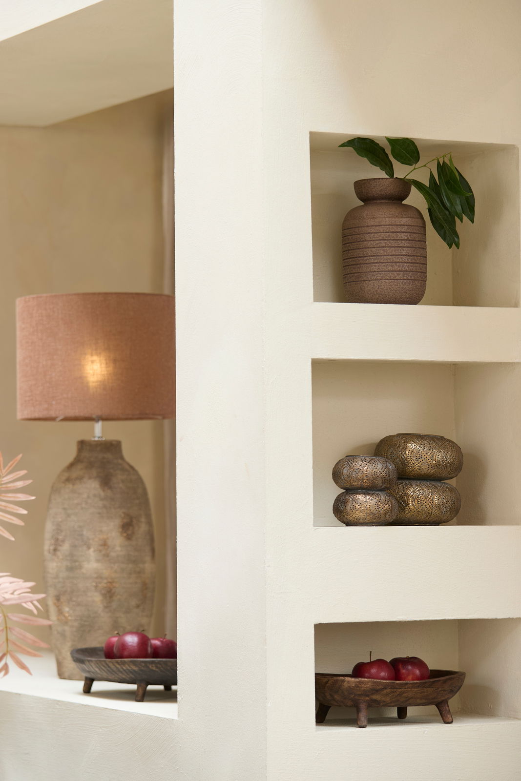 Ein stilvolles Windlicht in einem Regal, umgeben von dekorativen Elementen wie einer Vase mit Blättern und einer Tischlampe. Perspektive von vorne.