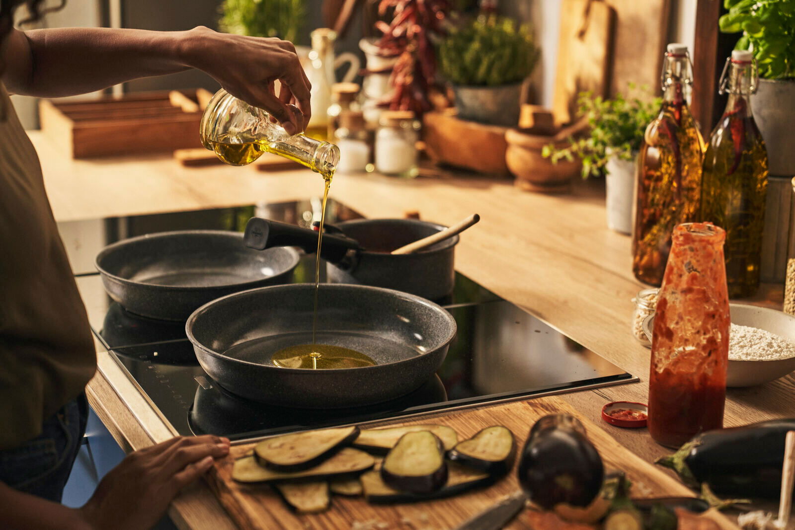 BALLARINI Pfannenset 2-tlg. Person gießt Öl in eine Pfanne auf einem Herd in einer gemütlichen Küche, seitliche Perspektive.
