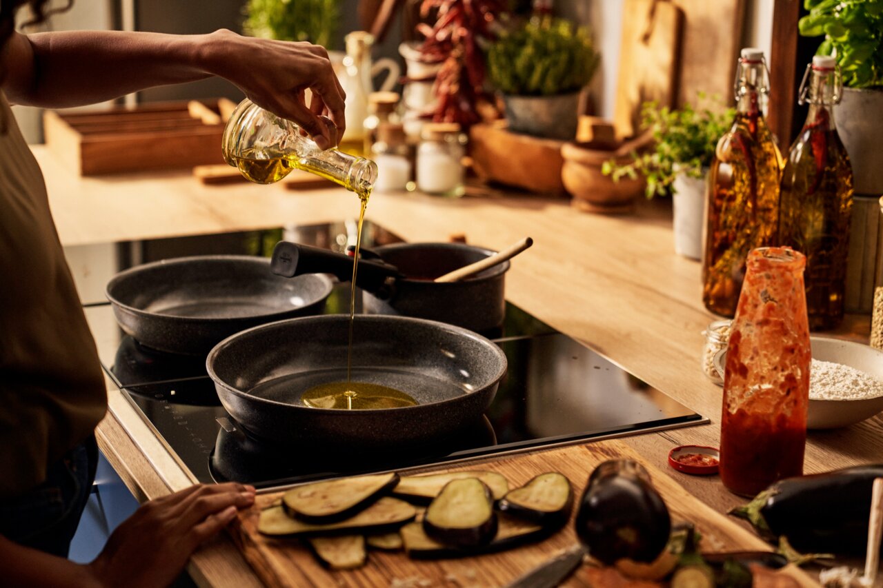 Person gießt Öl in eine Pfanne auf einem Herd in einer gemütlichen Küche, seitliche Perspektive.