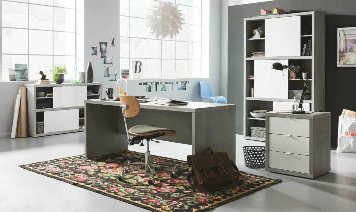 Moderner Schreibtisch in einem hellen Büro mit großem Fenster, umgeben von Regalen und einem bunten Teppich, aus seitlicher Perspektive.