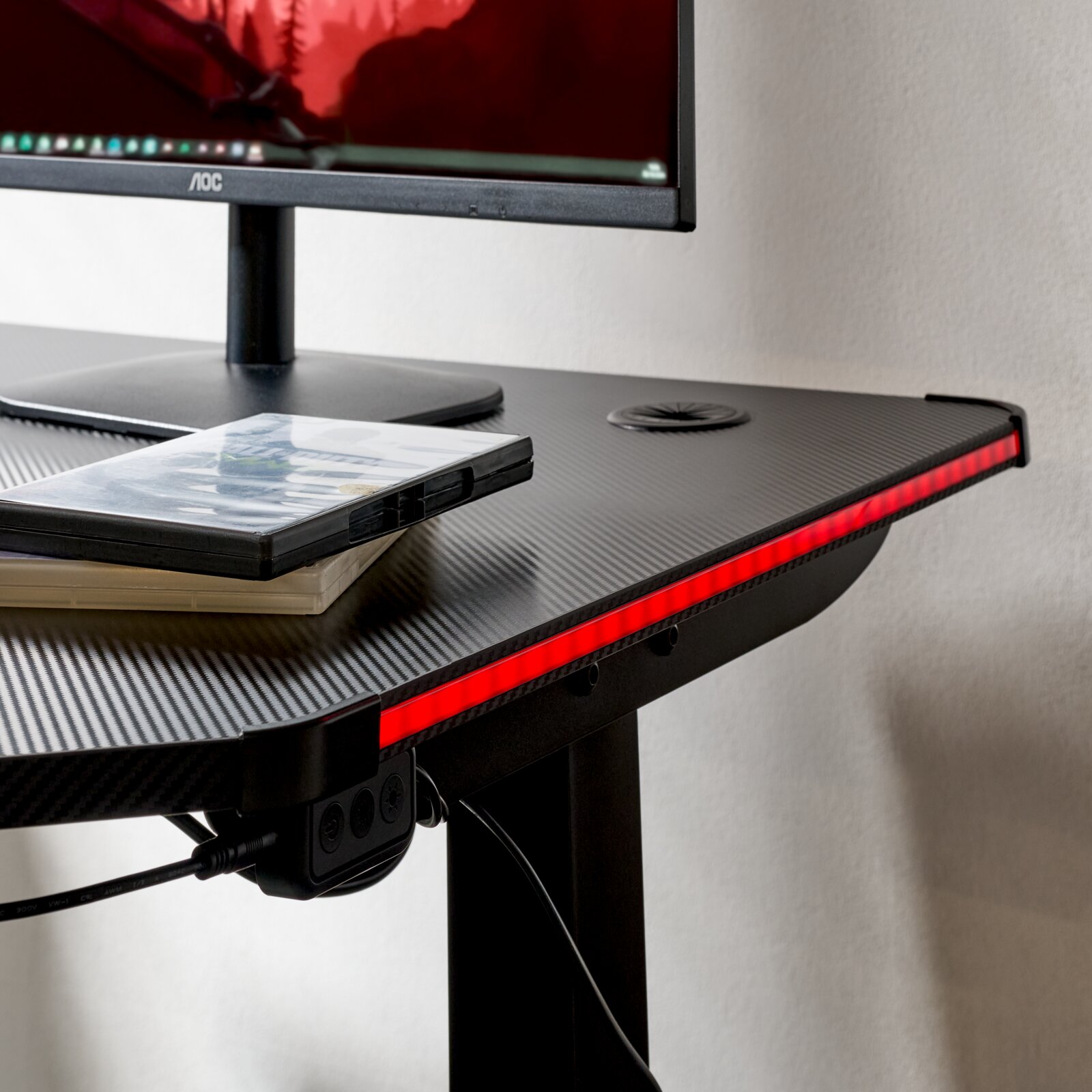 Gaming-Schreibtisch mit Carbon-Optik und roter LED-Kantenbeleuchtung, Monitor und Spielehüllen auf der Tischplatte, Ansicht schräg von vorne rechts (Nahaufnahme der rechten Vorderkante)