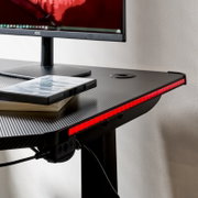 Gaming-Schreibtisch mit Carbon-Optik und roter LED-Kantenbeleuchtung, Monitor und Spielehüllen auf der Tischplatte, Ansicht schräg von vorne rechts (Nahaufnahme der rechten Vorderkante)