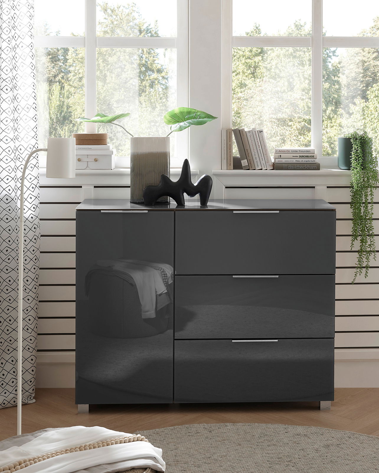 Moderne schwarze Kommode mit Hochglanzoberfläche, frontal fotografiert, dekoriert mit Pflanzen und Büchern vor einem Fenster.