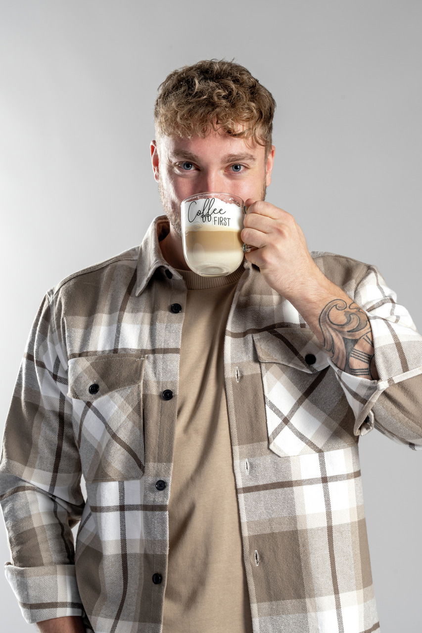 Mann in kariertem Hemd hält eine Tasse mit der Aufschrift 'Coffee FIRST' in der Hand, frontal aufgenommen.