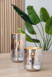 Zwei elegante Fink Windlichter BRIANO aus Metall mit geometrischem Muster, auf einem Holztisch stehend, im Hintergrund Pflanzen und Holzvertäfelung, seitliche Perspektive.