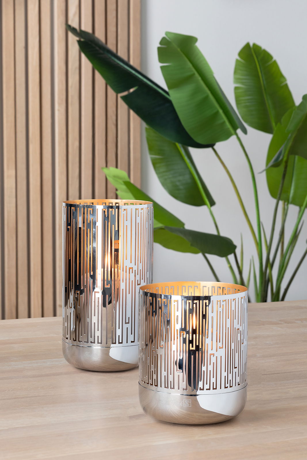 Zwei elegante Fink Windlichter BRIANO aus Metall mit geometrischem Muster, auf einem Holztisch stehend, im Hintergrund Pflanzen und Holzvertäfelung, seitliche Perspektive.