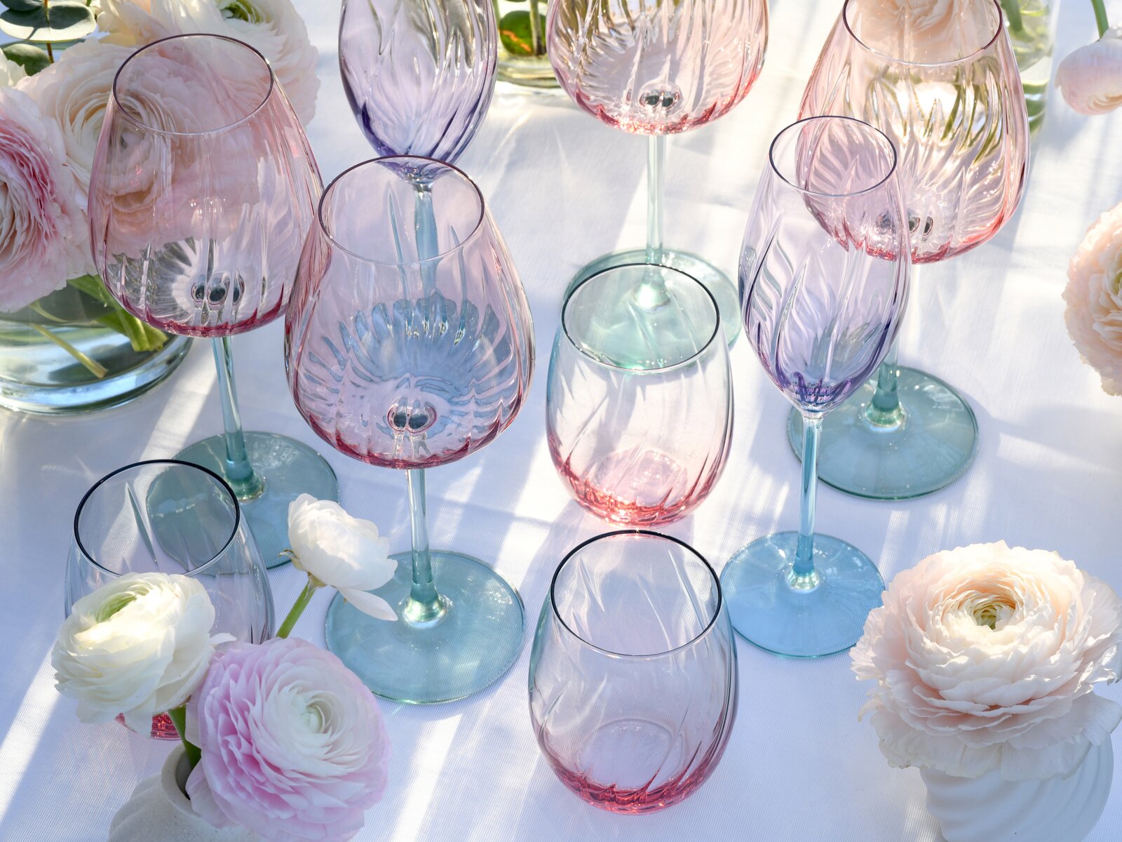 Schräg von oben fotografiertes Arrangement aus farbigen, strukturierten Weingläsern und Sektflöten aus Glas in Rosa- und Lilatönen mit türkisfarbenen Stielen auf weißer Tischdecke, dekoriert mit zarten Ranunkeln.