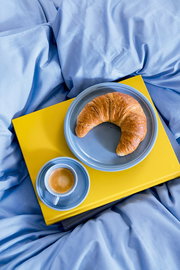Blauer Speiseteller mit Croissant und Espressotasse auf gelbem Tablett, von oben fotografiert, auf blauer Bettwäsche.