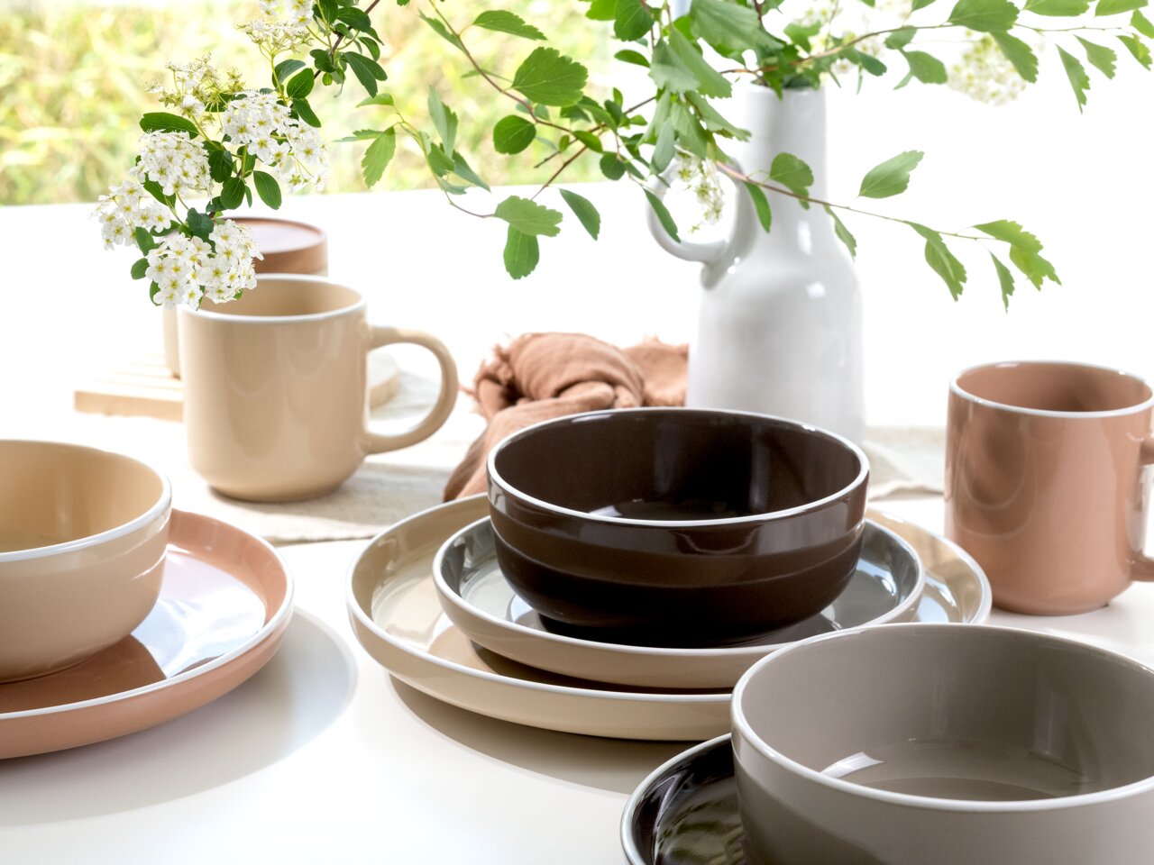 Kombiservice 16-tlg. in verschiedenen Erdtönen, bestehend aus Tellern, Schalen und Tassen, auf einem Tisch mit Blumenarrangement, seitliche Perspektive.
