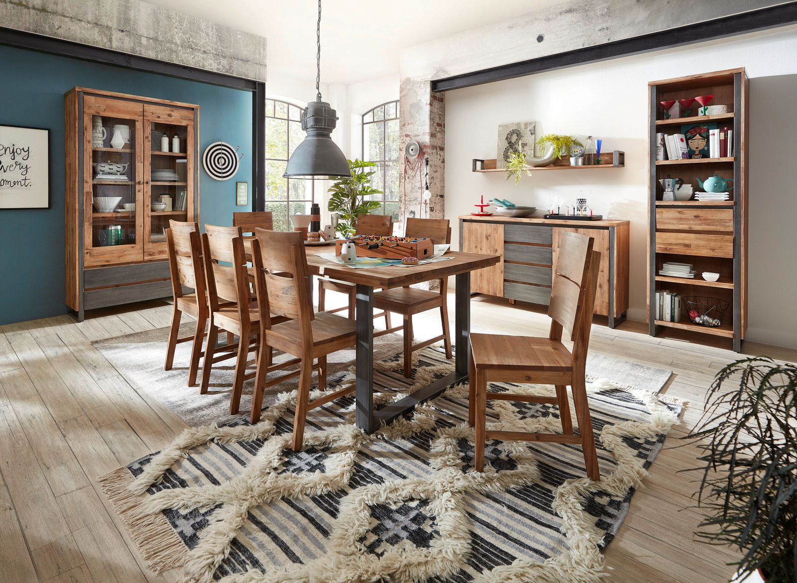 Esszimmer mit Holzmöbeln im rustikalen Stil, bestehend aus einem Esstisch, sechs Stühlen, einer Vitrine, einem Sideboard und einem Bücherregal. Die Perspektive zeigt den Raum von schräg vorne, mit einem Teppich auf dem Holzboden und großen Fenstern im Hin