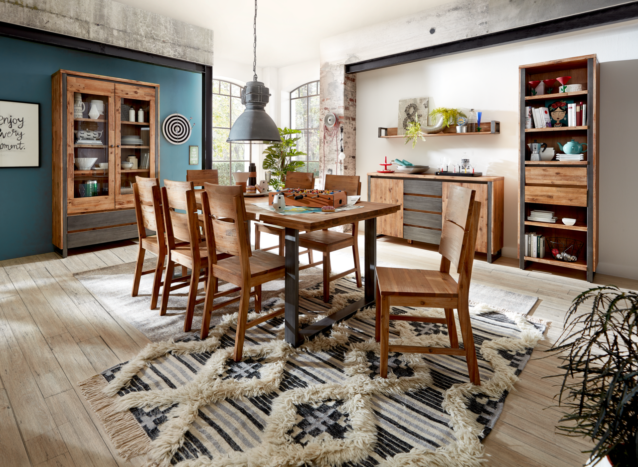 Landscape Stuhl I BALI Esszimmer mit Holzmöbeln im rustikalen Stil, bestehend aus einem Esstisch, sechs Stühlen, einer Vitrine, einem Sideboard und einem Bücherregal. Die Perspektive zeigt den Raum von schräg vorne, mit einem Teppich auf dem Holzboden und großen Fenstern im Hin