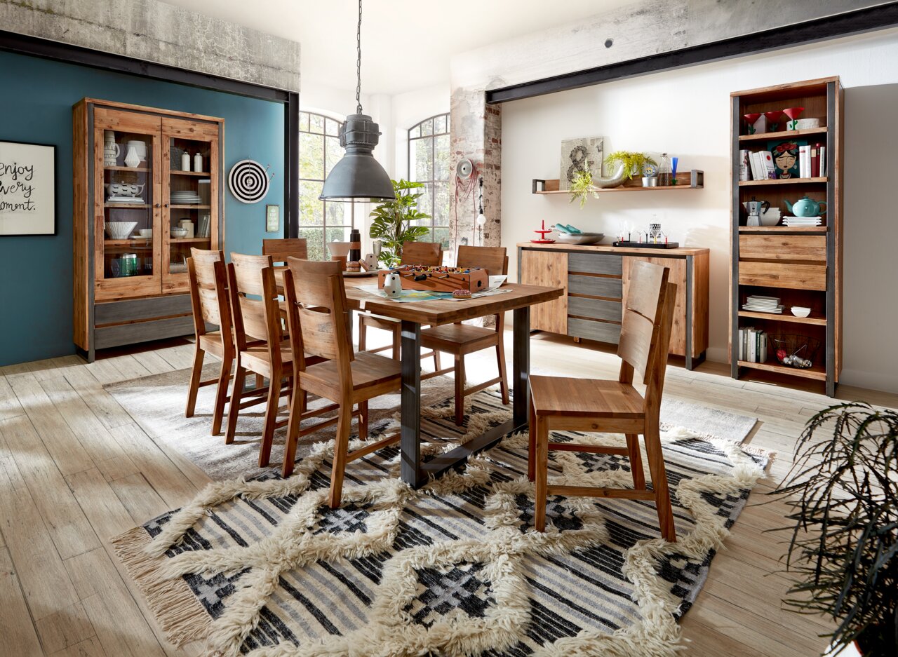 Esszimmer mit Holzmöbeln im rustikalen Stil, bestehend aus einem Esstisch, sechs Stühlen, einer Vitrine, einem Sideboard und einem Bücherregal. Die Perspektive zeigt den Raum von schräg vorne, mit einem Teppich auf dem Holzboden und großen Fenstern im Hin