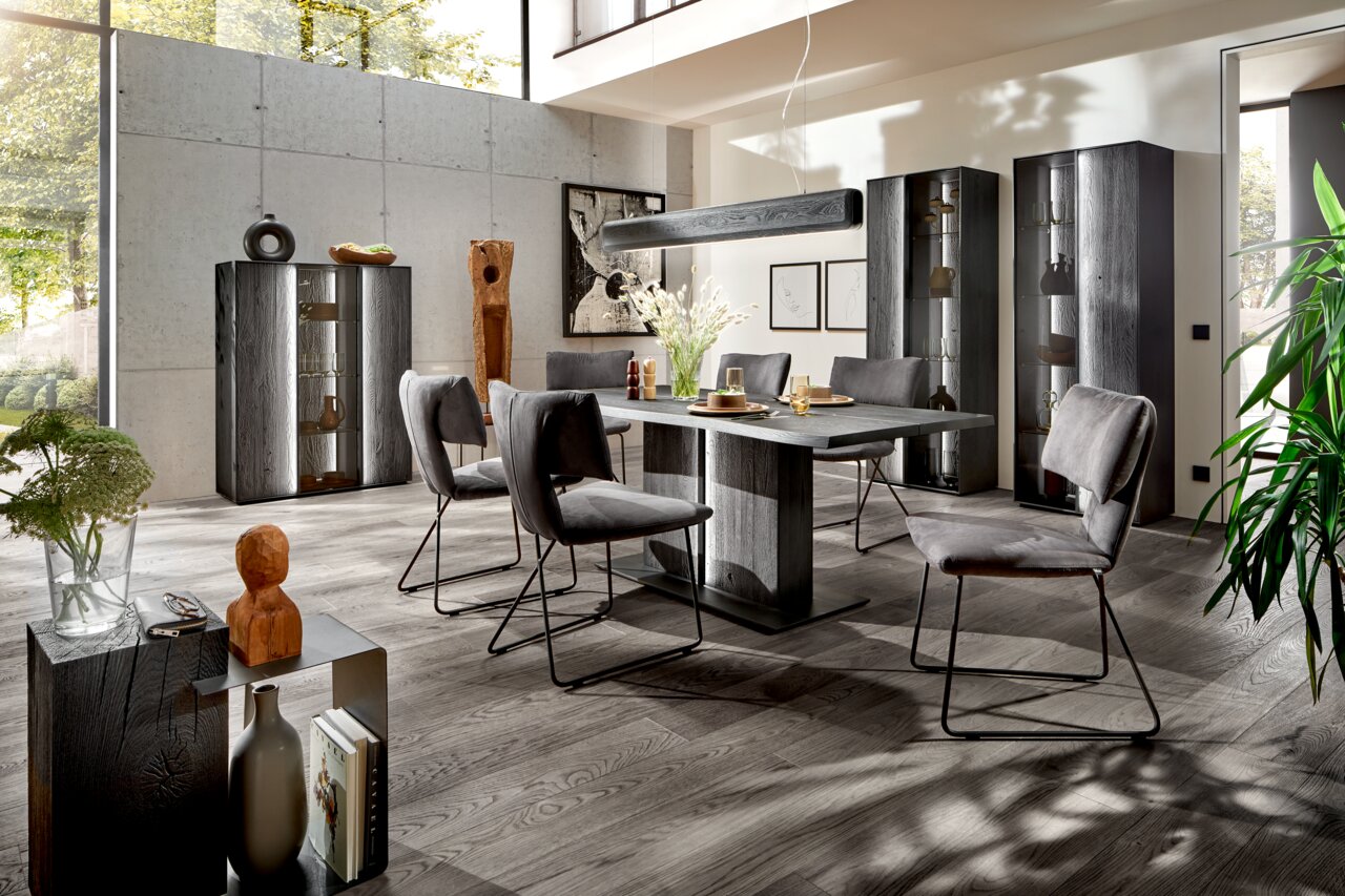 Moderne Esszimmer-Einrichtung mit grauem Esstisch und Stühlen, umgeben von Vitrinen und Dekorationen in einem hellen Raum mit großen Fenstern, seitliche Perspektive.