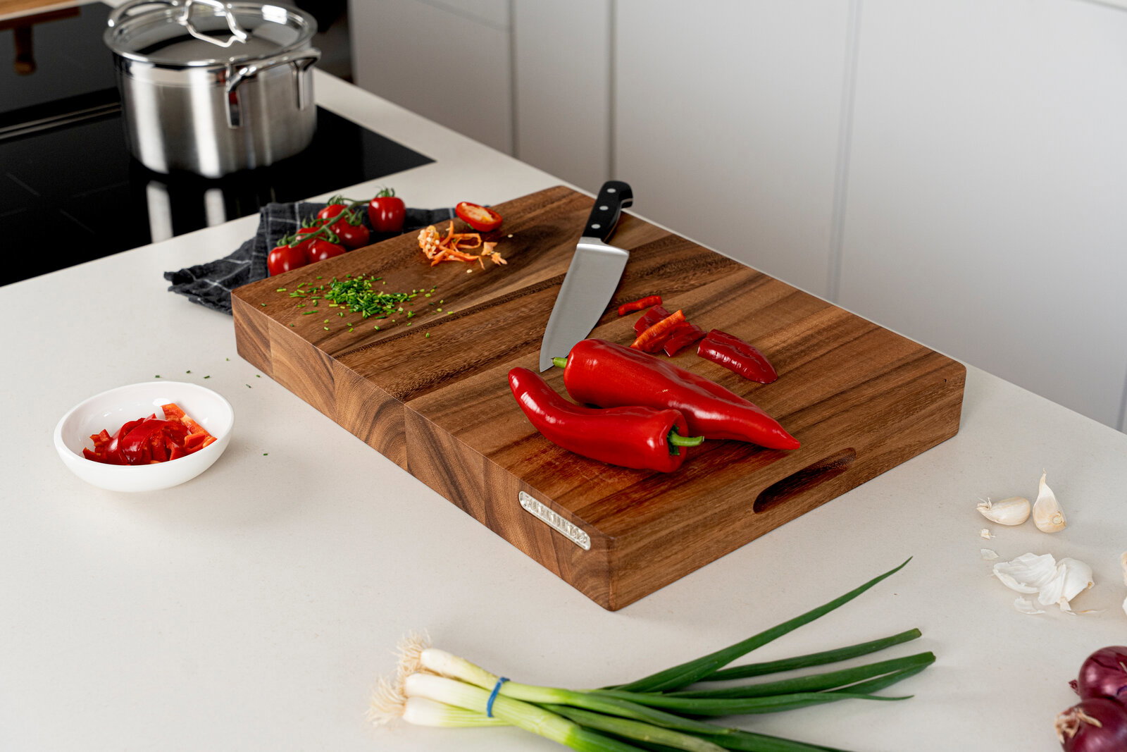 Großes, massives Holz-Schneidebrett auf heller Küchenarbeitsplatte mit Kochmesser, roten Paprika/Chilis und gehackten Kräutern; Perspektive: leicht von oben aus schrägem Winkel.