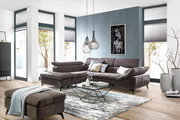 Ecksofa in modernem Wohnzimmer, Perspektive von vorne links, mit grauem Stoffbezug, dekorativen Kissen und einem passenden Hocker.