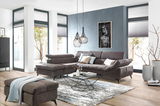 Ecksofa in modernem Wohnzimmer, Perspektive von vorne links, mit grauem Stoffbezug, dekorativen Kissen und einem passenden Hocker.
