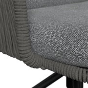 Detailfoto van een grijs kussen met een gestructureerd oppervlak en een rondomlopend gevlochten koord op een rotan-/vlechtmeubel, vanuit een schuin perspectief van bovenaf op de zitrand