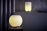 Zwei moderne, kugelförmige Tischlampen aus Messing mit warmem Licht, eine im Vordergrund und eine im Hintergrund auf einem Regal platziert.