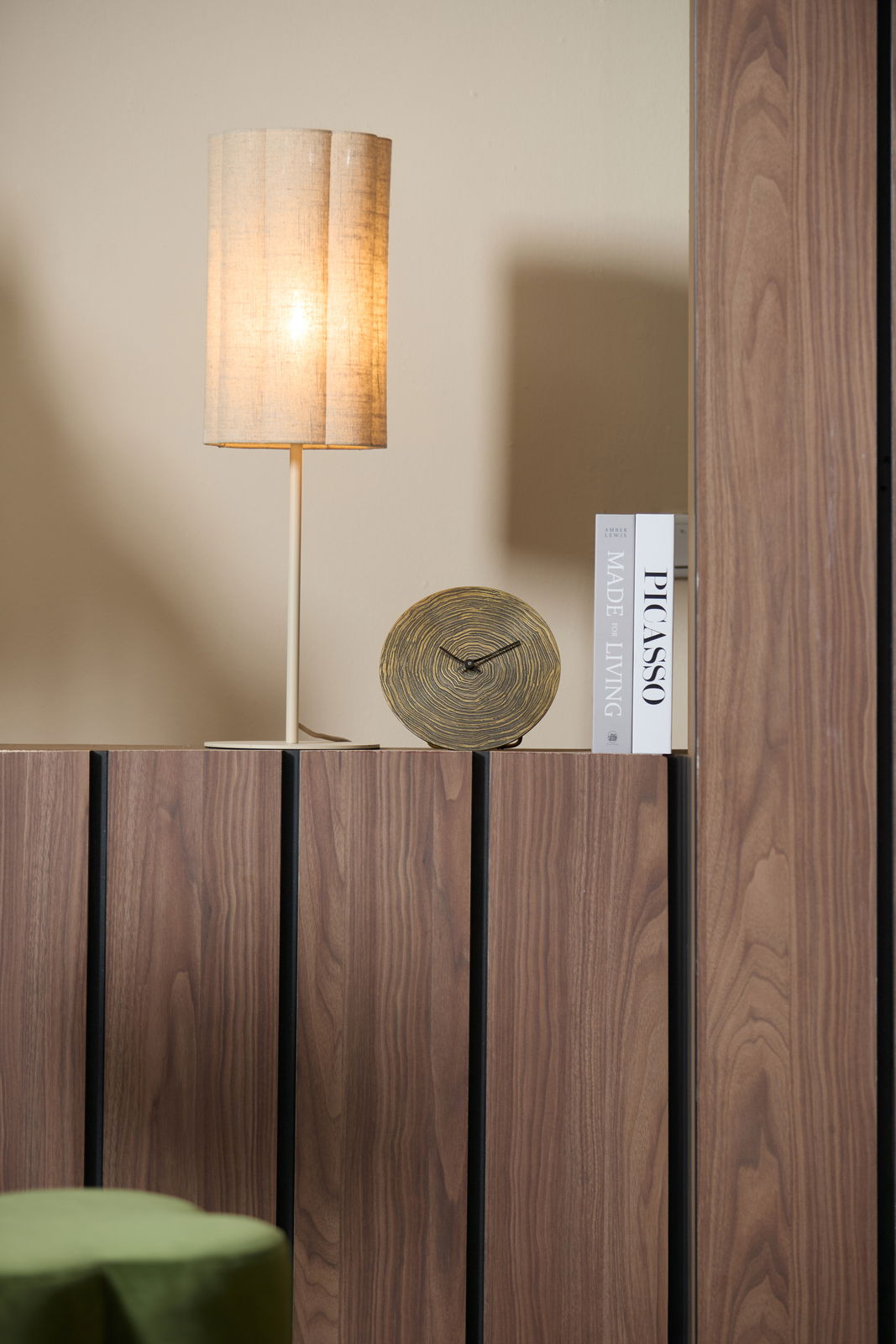 Runde Tischuhr mit Holzoptik, frontal auf einem Holzregal neben einer beleuchteten Tischlampe und Büchern platziert.