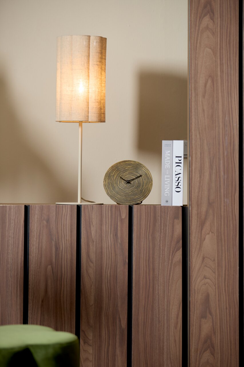 Runde Tischuhr mit Holzoptik, frontal auf einem Holzregal neben einer beleuchteten Tischlampe und Büchern platziert.