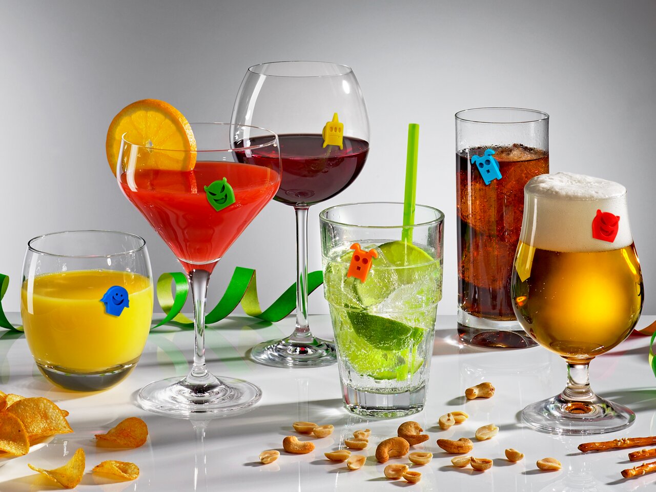 Verschiedene Gläser mit bunten Glasmarkern in Form von Geistern, gefüllt mit verschiedenen Getränken, auf einer weißen Oberfläche. Perspektive: Frontalansicht.