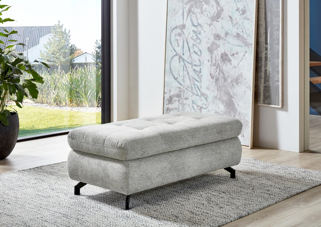 Grauer XXL-Hocker mit schwarzem Metallgestell in einem modernen Wohnzimmer, seitliche Perspektive.