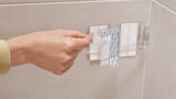 Nahaufnahme einer Hand, die ein Klebepad von einer transparenten Halterung an einer Badezimmerwand abzieht.