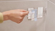 Nahaufnahme einer Hand, die ein Klebepad von einer transparenten Halterung an einer Badezimmerwand abzieht.