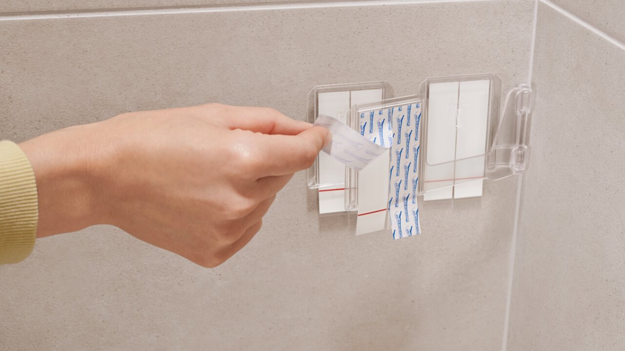 Nahaufnahme einer Hand, die ein Klebepad von einer transparenten Halterung an einer Badezimmerwand abzieht.