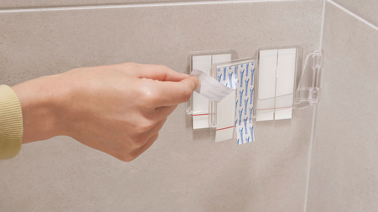 Nahaufnahme einer Hand, die ein Klebepad von einer transparenten Halterung an einer Badezimmerwand abzieht.