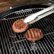 Barbecue-Grillzange aus Edelstahl greift einen Burger auf einem Grillrost, seitliche Perspektive