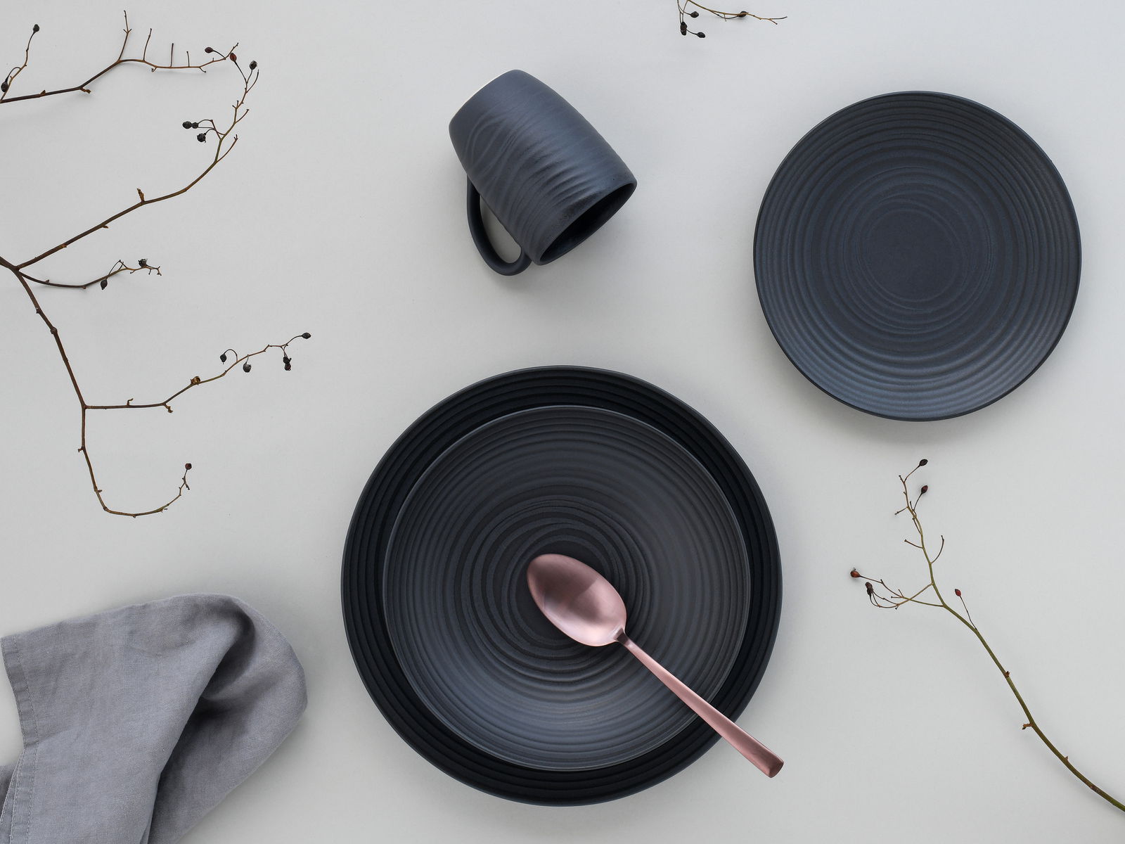 Draufsicht auf ein elegantes, dunkelgraues Kombiservice-Set mit Teller, Tasse und Löffel, dekoriert mit Zweigen und Stoffserviette.