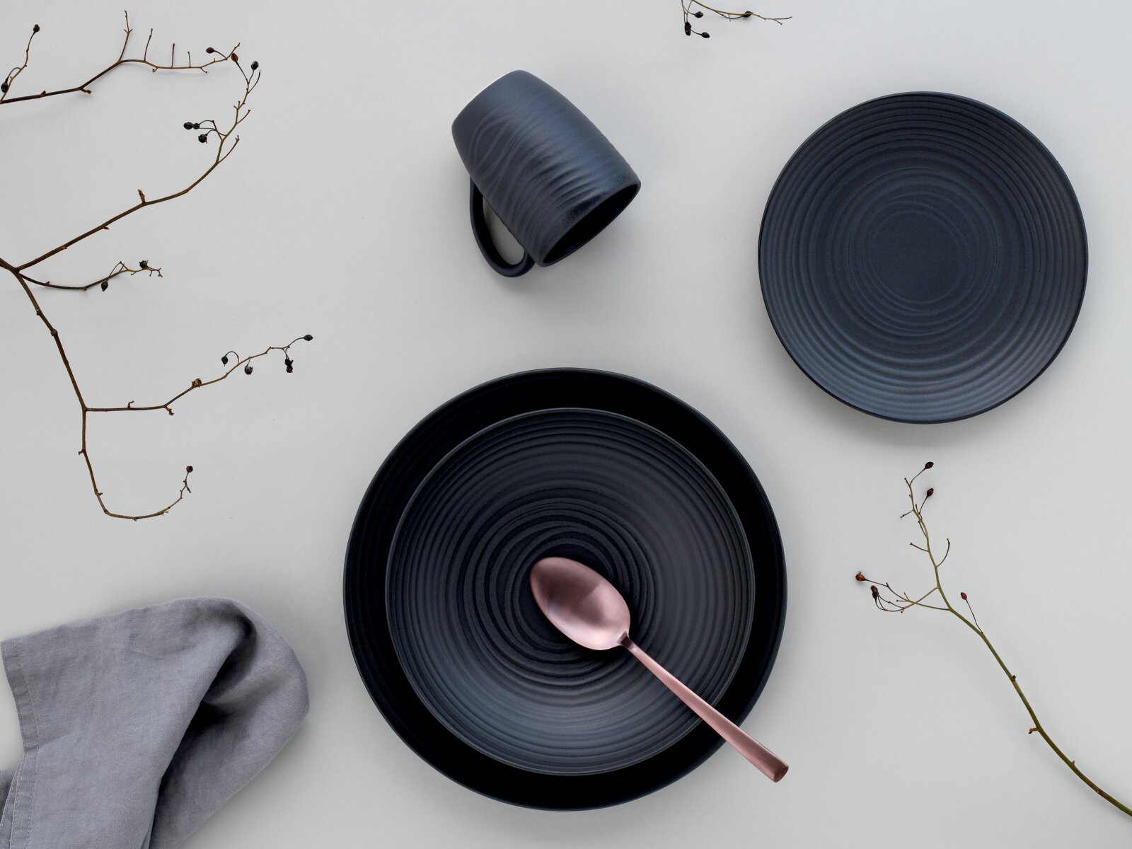 Draufsicht auf ein elegantes, dunkelgraues Kombiservice-Set mit Teller, Tasse und Löffel, dekoriert mit Zweigen und Stoffserviette.