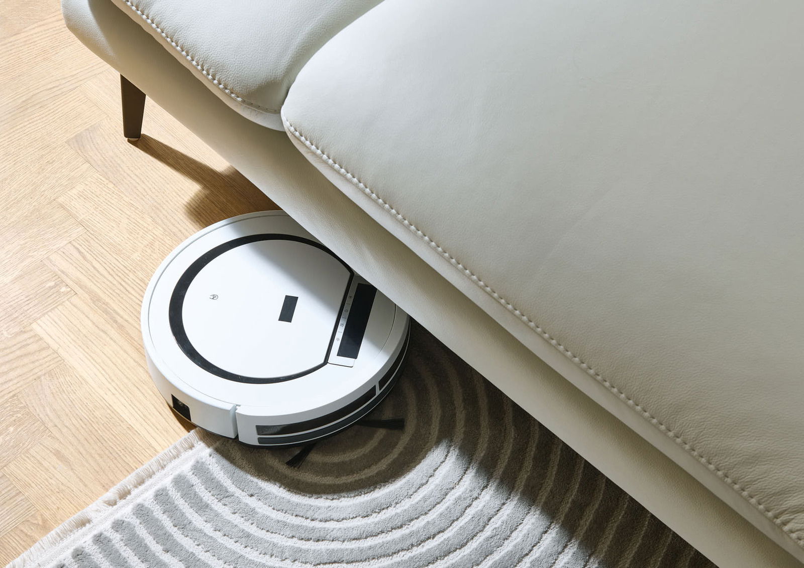 Ein weißer Saugroboter reinigt den Boden unter einem hellen Sofa aus der Vogelperspektive.