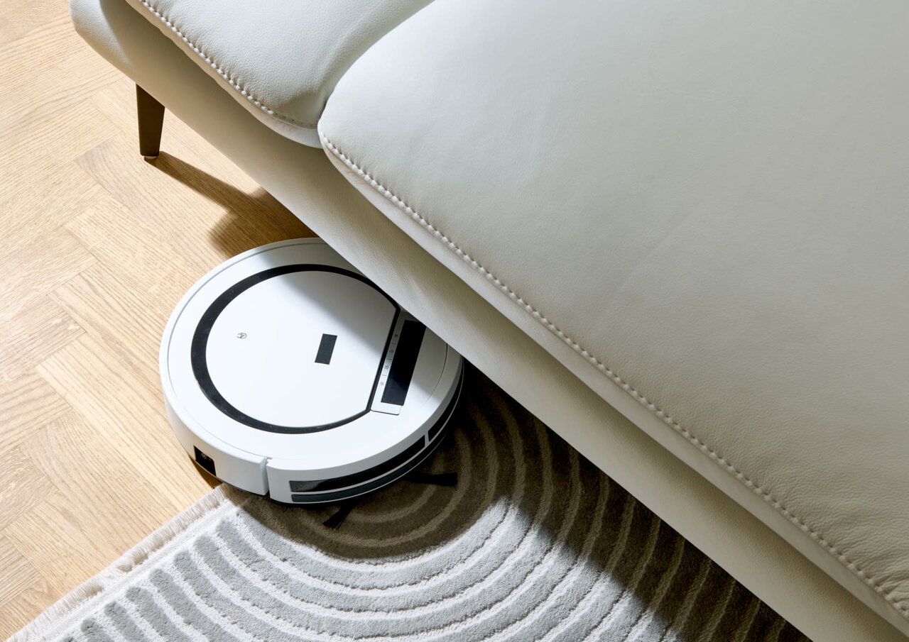 Ein weißer Saugroboter reinigt den Boden unter einem hellen Sofa aus der Vogelperspektive.