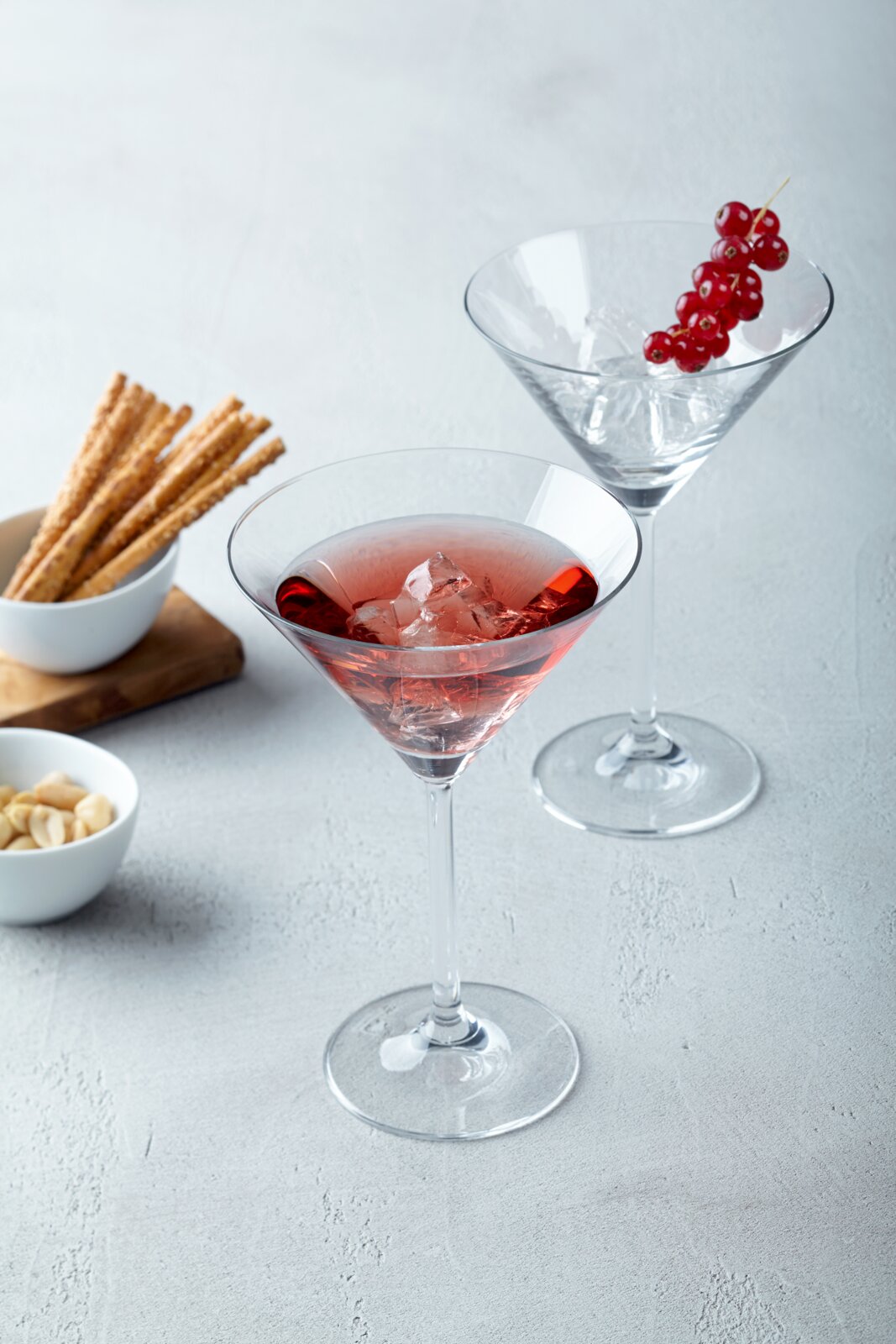 Zwei elegante Martini-Cocktailgläser aus klarem Glas auf hellgrauer Oberfläche, vorn ein roséfarbener Drink mit Eiswürfeln, hinten ein klares Getränk mit roten Johannisbeeren; Perspektive: leicht von oben in schräger Frontansicht