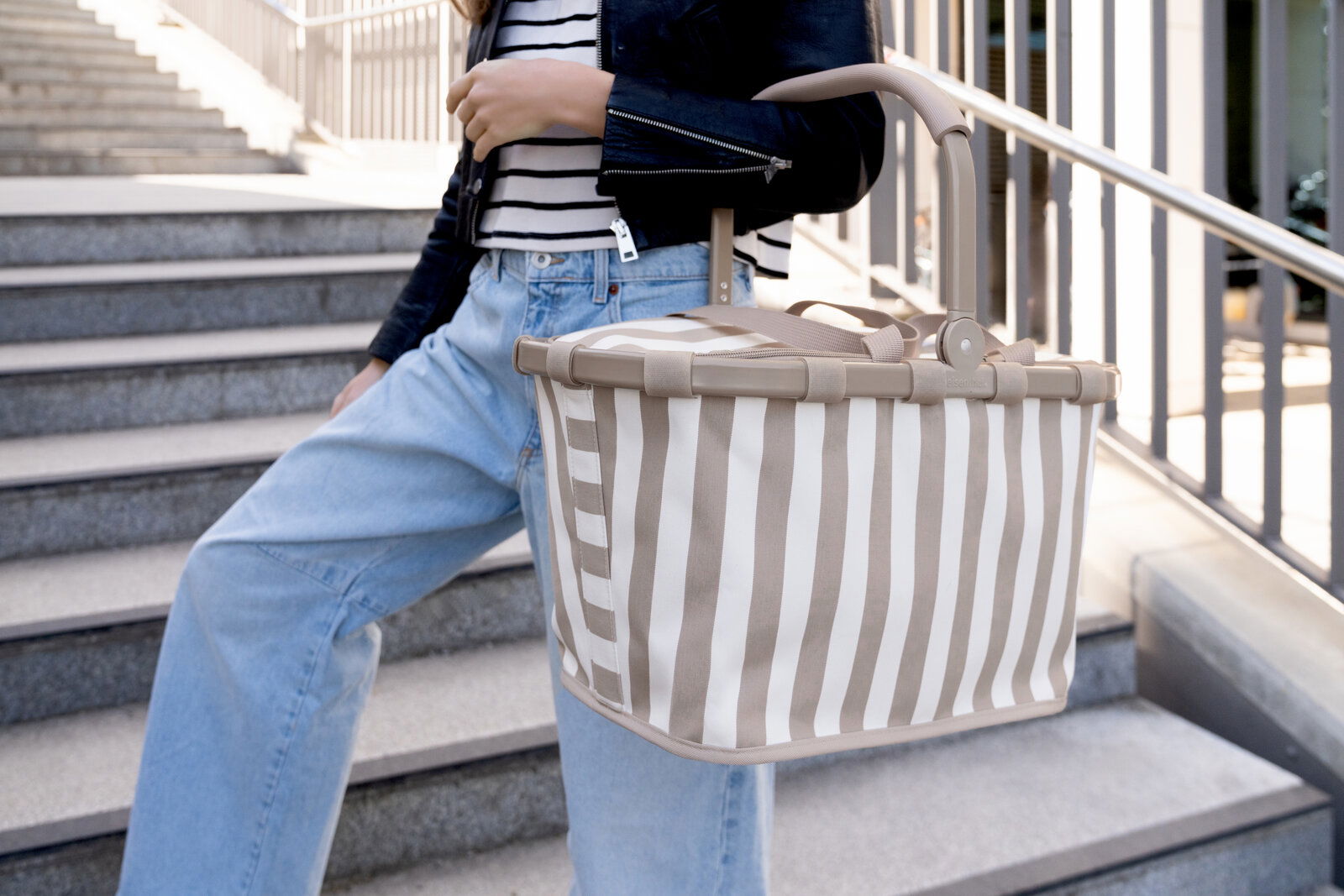 Beige-weiß gestreifter Einkaufskorb mit klappbarem Tragebügel, seitlich in leichter Untersicht fotografiert, getragen von einer Person auf einer Treppe im Außenbereich.
