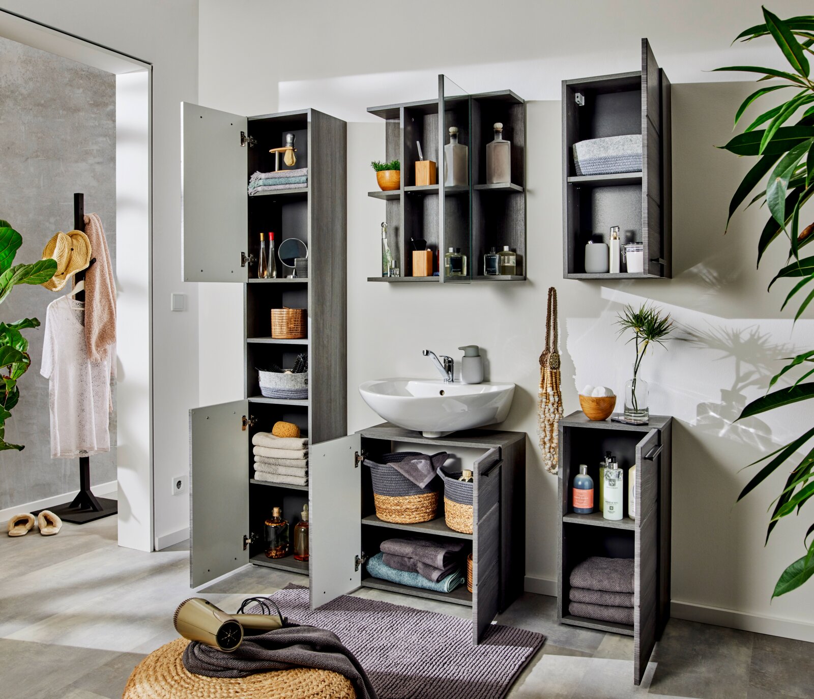 Modernes Badezimmermöbel-Set in grauer Holzoptik mit Hochschrank, Hängeschränken und Waschtischunterschrank mit Aufsatzwaschbecken, Türen teils geöffnet; Frontansicht leicht schräg von links.