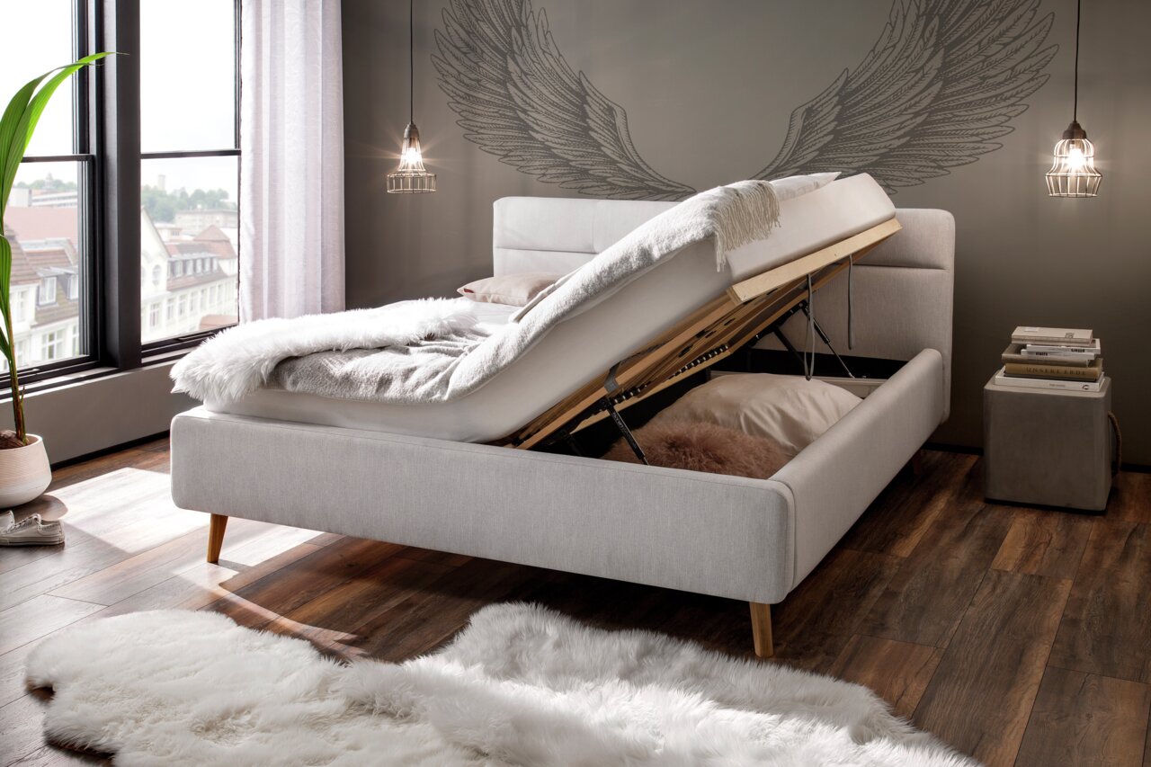 Modern gestoffeerd bed met opbergruimte, gezien vanaf de zijkant, in een stijlvol ingerichte slaapkamer met grote ramen en een houten vloer.